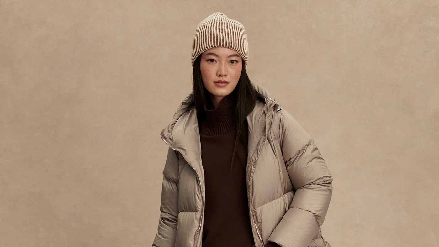 Woman wearing a beige puffer jacket and knit hat against a beige background